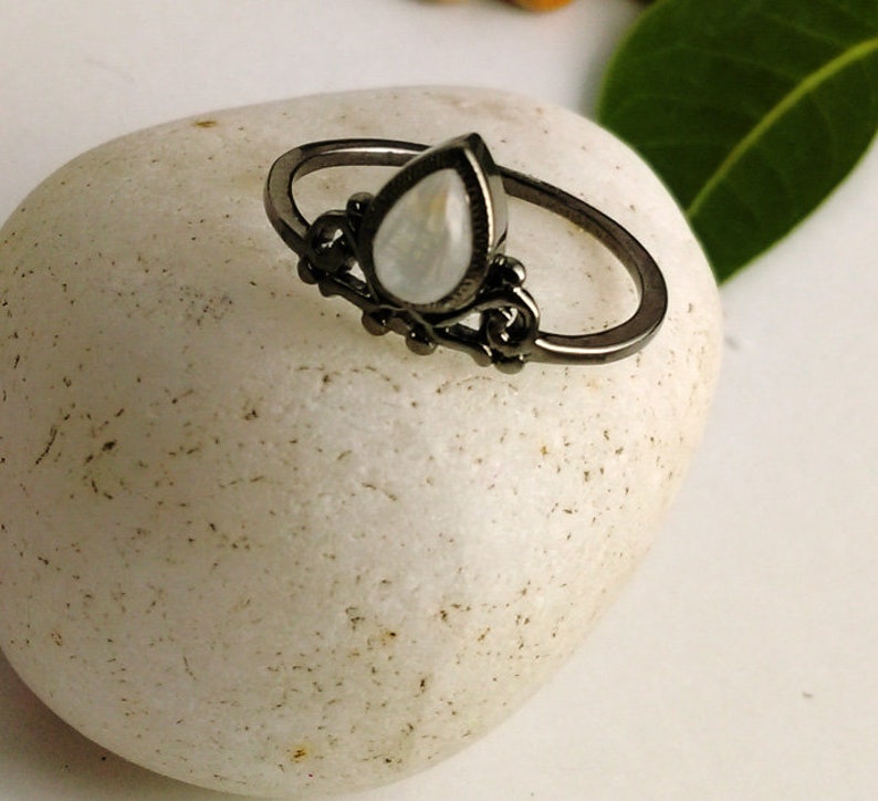 May include: A black metal ring with a pear-shaped, iridescent gemstone. The ring features ornate detailing around the gemstone and along the band. The ring is set on a white, textured surface, showcasing the jewelry's design and color.