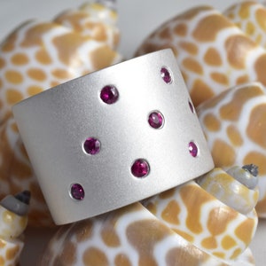 May include: A wide, silver-toned cuff bracelet with a matte finish. The bracelet is adorned with several small, round, deep pink gemstones evenly spaced across its surface. The background features several seashells with a tan and white spotted pattern.