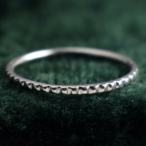 May include: A silver ring with a textured, dotted pattern around the band. The ring is thin and delicate, set against a dark green background. The ring is likely made of sterling silver.
