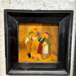 Könnte beinhalten: Gerahmtes Gemälde, das eine Volkszene mit drei Figuren und einem Mann in traditioneller Kleidung zeigt. Das Gemälde hat einen schwarzen Rahmen und eine warme Farbpalette, wobei die Figuren farbenfrohe Kleidung tragen.
