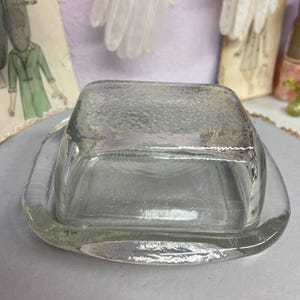 Könnte beinhalten: Ein klarer, quadratischer Butterbehälter aus Glas mit strukturierter Oberfläche. Die Schale hat einen passenden Deckel und einen abgerundeten Boden. Der Butterbehälter ist für eine Standardbuttergröße konzipiert.