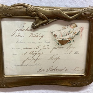 Könnte beinhalten: Ein antiker, messingfarbener Bilderrahmen mit Jugendstil-Design. Der Rahmen zeigt eine liegende Figur oben und aufwendige florale Details. Im Inneren befindet sich ein gedrucktes Bild eines Babys in einem Korb und handschriftlicher Text. Der Rahmen misst ca. 18 x 13 cm.