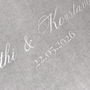 May include: A close-up shot of grey fabric with white cursive text. The text reads "thi & Konstanti" and the date "22.05.2026". The word "MARISEY" is at the bottom. The font is elegant and delicate.