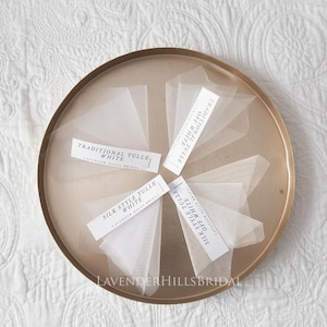 Peut inclure: Cinq échantillons de tulle sur un plateau doré. Les échantillons sont étiquetés "Tulle Traditionnel Blanc", "Tulle Style Soie Blanc", "Tulle Traditionnel Blanc Cassé", et "Tulle Style Soie Blanc Cassé". Le plateau est étiqueté "Lavender Hills Bridal".