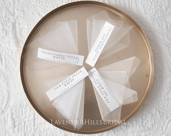 May include: Five swatches of tulle fabric on a gold tray. The swatches are labeled "Traditional Tulle White", "Silk Style Tulle White", "Traditional Tulle Off White", and "Silk Style Tulle Off White". The tray is labeled "Lavender Hills Bridal".