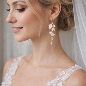 May include: Gold-coloured earrings featuring white floral drops and small dangling pearls. The earrings have a delicate design with layered petals, suitable for bridal or special events. A sophisticated accessory.