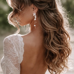 Peut inclure: Dos et épaule d'une femme vêtue d'une robe en dentelle blanche et de boucles d'oreilles florales. Les boucles d'oreilles sont composées de fleurs blanches délicates et de petites perles. Cheveux longs, ondulés et châtain clair. Le mot "MARISEY" est visible.