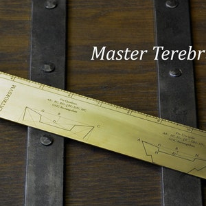 Könnte beinhalten: Ein Messstab aus Messing mit geätzten Diagrammen und Text, beschriftet mit "Master Terebrus". Der Messstab liegt auf einer Holzoberfläche mit Metallbändern. Er zeigt geometrische Formen und Messmarkierungen.