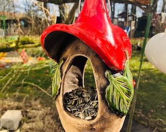 Op de afbeelding: Een hangende vogelvoeder met een rood, conisch dak en een bruine, keramische behuizing. De voeder is gevuld met zonnebloempitten en versierd met groene keramische bladeren. Het hangt aan een tak.