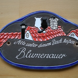 Könnte beinhalten: Ein schwarz-weißes Keramik-Hausschild mit roten Dachziegeln und zwei Katzen, die auf dem Dach sitzen. Das Schild trägt die Aufschrift "Alle unter diesem Dach heißen Blumenauer".