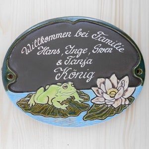 Könnte beinhalten: Ein schwarzes ovales Schild aus Keramik mit weißer Aufschrift "Willkommen bei Familie Hans, Inge, Sven & Tanja König". Das Schild hat einen grünen Frosch, der auf einem Seerosenblatt sitzt, und eine weiße Seerose mit gelber Mitte.