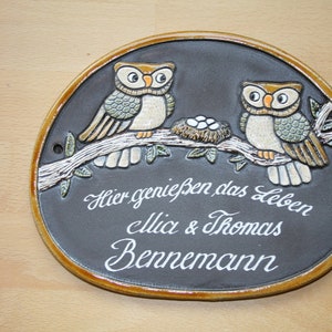 Könnte beinhalten: Ovale Keramikplakette mit zwei Eulen auf einem Ast, einem Nest mit Eiern und dem Text "Hier genießen das Leben, Mia & Thomas Bennemann". Sie hat einen dunkelgrauen Hintergrund und einen braunen Rand.