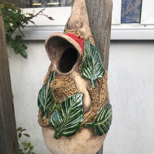 Könnte beinhalten: Ein Keramik-Vogelhaus in Tropfenform mit einem roten Dach und grünen Blättern um die Basis. Das Vogelhaus ist an einem Holzpfosten befestigt.