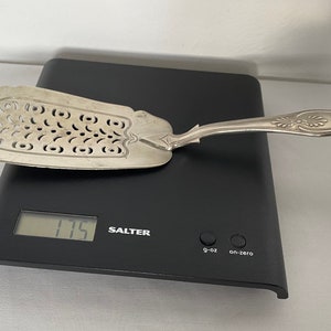 May include: A silver fish server with an ornate handle and a pierced, curved blade. The server rests on a black digital scale, showing a weight of 175. The scale displays the weight in g and oz.