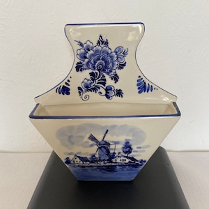 May include: A white ceramic wall pocket with blue floral and windmill designs. The top has a handle and a large blue floral pattern. The bottom features a landscape scene with a windmill, houses, and water. The wall pocket is on a black scale.