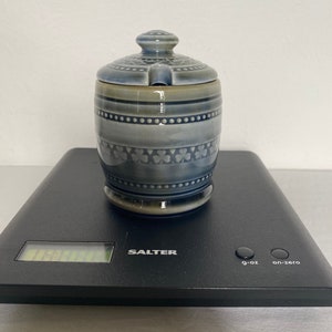 May include: A blue and grey ceramic sugar bowl with a lid. The bowl has a cylindrical shape with decorative patterns, including dots and clover-like shapes. The lid has a small handle and a slot for a spoon. The bowl is on a black scale.