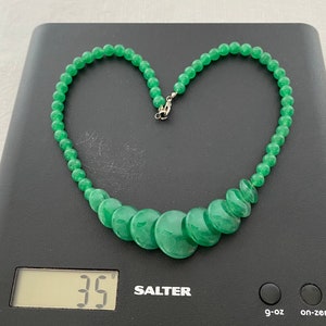 May include: A bright green jade necklace, featuring small round beads and larger, flat, disc-shaped beads. The necklace is on a black digital scale, which reads 35 grams. The clasp is silver-coloured.