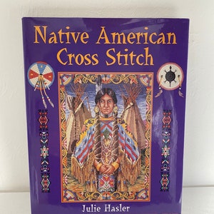 May include: A book titled "Native American Cross Stitch" with a purple cover. The cover displays a cross-stitch image of a Native American woman in traditional dress, with teepees in the background. The author's name, Julie Hasler, is printed at the bottom.