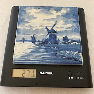 May include: A black digital kitchen scale with a blue and white tile top depicting a windmill scene. The display shows 271 g. The scale has g-oz and on-zero buttons. The brand name "Salter" is visible.