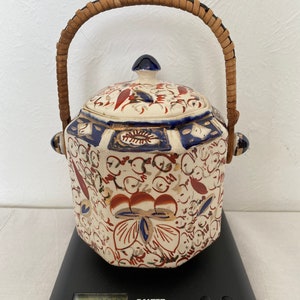 May include: A ceramic jar with a lid and a woven handle. The jar has a cream base with red, blue, and gold floral patterns. The jar is on a black digital scale. The scale displays a weight.