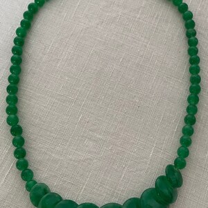 May include: A green beaded necklace with a silver clasp. The necklace features small, round beads that gradually increase in size towards the centre, creating a graduated design. The beads have a glossy finish.