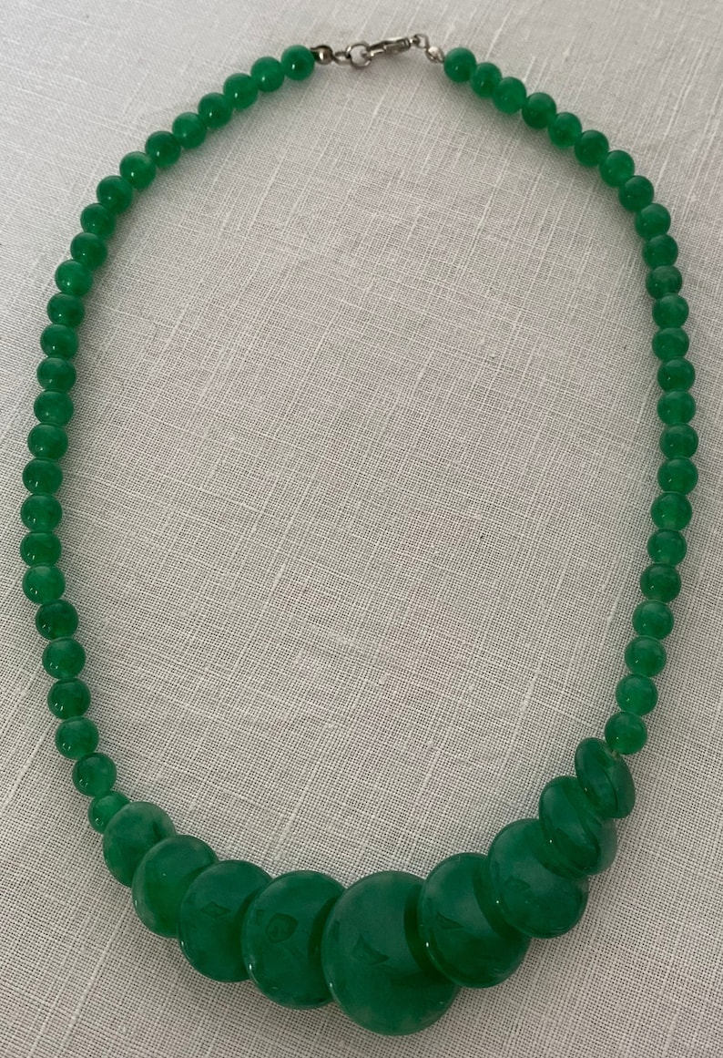 May include: A green beaded necklace with a silver clasp. The necklace features small, round beads that gradually increase in size towards the centre, creating a graduated design. The beads have a glossy finish.