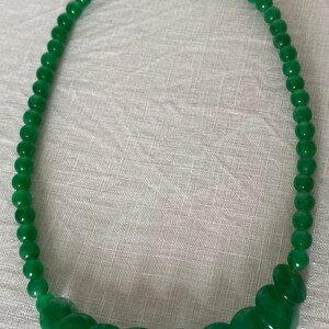 May include: A green beaded necklace featuring a mix of round and flat beads. The necklace has a silver clasp and is shown on a white surface. The beads are a vibrant, solid green colour.