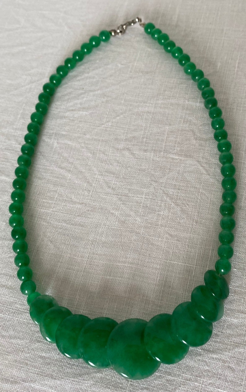 May include: A green beaded necklace featuring a mix of round and flat beads. The necklace has a silver clasp and is shown on a white surface. The beads are a vibrant, solid green colour.