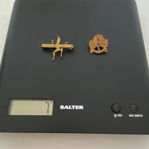 May include: Two vintage gold-coloured pins on a black scale. One pin is a stylized figure, the other is a trefoil design. The scale displays a weight of 7 g-oz. The scale is labelled "Salter".