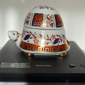May include: A white ceramic turtle-shaped trinket box with a lid. The box is decorated with orange, blue, and gold floral patterns. The turtle has a smiling face and gold accents. The box is on a black surface. The measurements are in centimetres.