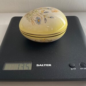 May include: A yellow, egg-shaped trinket box with a floral design in blue, yellow, and green. The box has a gold trim and is sitting on a black scale. The scale reads 135.