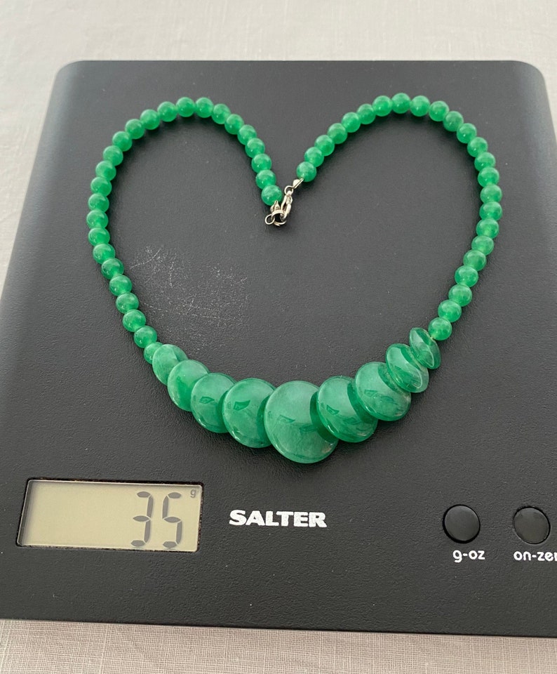 May include: A bright green jade necklace, featuring small round beads and larger, flat, disc-shaped beads. The necklace is on a black digital scale, which reads 35 grams. The clasp is silver-coloured.