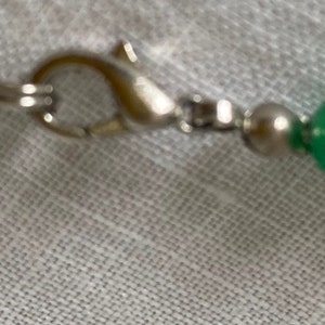 May include: Close-up of a green beaded necklace with a silver clasp. The beads are round and have a glossy finish. The necklace is strung on a silver chain.