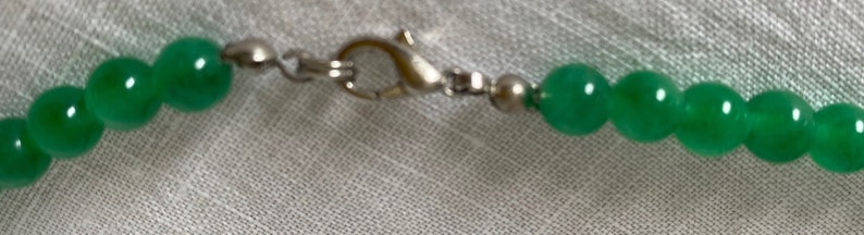 May include: Close-up of a green beaded necklace with a silver clasp. The beads are round and have a glossy finish. The necklace is strung on a silver chain.