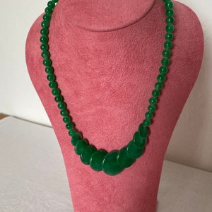 May include: A green jade necklace on a pink jewellery bust. The necklace has graduated round beads and flat, circular jade pieces. The jade has a polished finish, and the beads vary in size. The necklace is displayed on a pink bust.