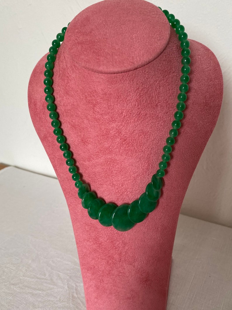 May include: A green jade necklace on a pink jewellery bust. The necklace has graduated round beads and flat, circular jade pieces. The jade has a polished finish, and the beads vary in size. The necklace is displayed on a pink bust.