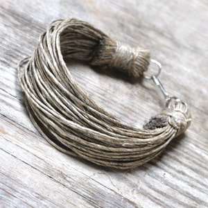 May include: A handmade bracelet crafted from tightly wound, natural-colored twine. The bracelet features a silver-tone clasp for closure. The twine is arranged in a circular shape, showcasing the texture and earthy tones of the material.