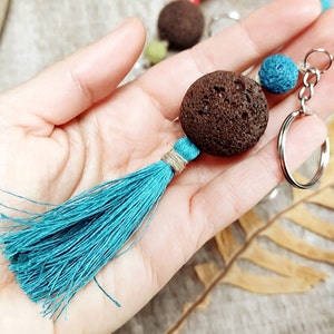 Könnte beinhalten: Ein türkisfarbener Quasten-Schlüsselanhänger mit einem braunen Lavastein-Anhänger und einem blauen Perlenakzent. Der Schlüsselanhänger ist an einem silbernen Schlüsselring befestigt.