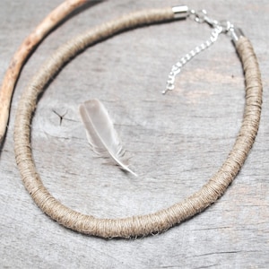 May include: A circular necklace featuring a thick cord wrapped in tan jute twine. The necklace includes a silver-toned metal chain extender with a lobster claw clasp and a single small, light gray feather resting inside the circular frame.
