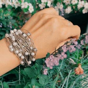 May include: A bracelet made of brown string and white pearls. The bracelet is wrapped around a person's wrist and is worn against a background of green leaves and pink flowers.