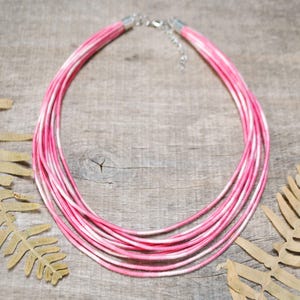 Può includere: Una collana a più fili con cordoncini rosa e bianchi. La collana ha una chiusura argentata e una catena regolabile. I cordoncini sono disposti in un design a strati, creando un accessorio audace. La collana è esposta su una superficie di legno.