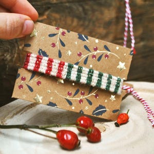Puede incluir: Una pulsera tejida con rayas rojas, blancas y verdes está sujeta a una etiqueta de regalo marrón decorada con estrellas y ramitas doradas. La etiqueta está sujeta por una mano. También se ven bayas rojas y una cuerda roja y blanca.