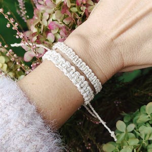 May include: Two white macrame bracelets worn on a wrist. The bracelets are made with a simple knot pattern and have a natural, earthy look.