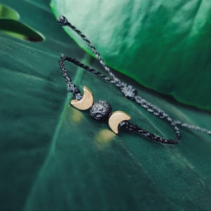 Może przedstawiać: Bransoletka ze sznurka w kolorze czarnym z trzema złotymi zawieszkami w kształcie półksiężyca i czarną koralikiem z lawy.