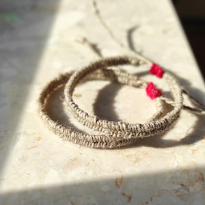 Op de afbeelding: Twee ronde armbanden van beige touw, met kleine rode accenten. De armbanden zijn gemaakt van natuurlijke vezels. De foto is genomen in natuurlijk licht, waardoor de textuur en het vakmanschap worden benadrukt.