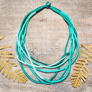 Può includere: Collana fatta a mano con più fili di corda turchese e azzurra. La collana è annodata in alto e poggia su una superficie di legno, con foglie secche sullo sfondo. Un gioiello di grande effetto.