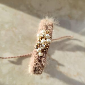 May include: A handmade, light brown crochet bracelet adorned with faux pearls and gold beads. The bracelet has a fluffy texture and a delicate, feminine design, perfect for adding a touch of elegance to any outfit. The bracelet is a unique piece of jewelry.