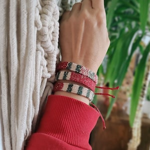 Puede incluir: Tres pulseras de amistad tejidas en colores rojo, blanco, verde y dorado. Las pulseras están apiladas en la muñeca de una persona.