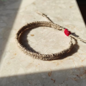 May include: Handmade bracelet made from natural jute twine. The bracelet has a woven design with a small red accent. The adjustable closure is made of the same jute material. A simple, earthy accessory.