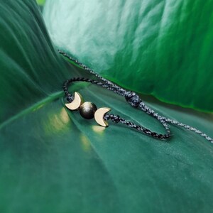 以下が含まれることがあります： 黒い紐のブレスレットに、金の三日月形のチャームと中央に黒いビーズが付いている。